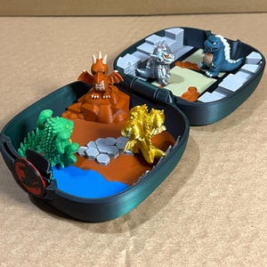 Könnte beinhalten: Ein offener, dunkelgrüner, aufklappbarer Behälter mit Miniatur-Dinosaurierfiguren. Der Behälter zeigt eine Landschaft mit einem blauen See, orangefarbenem Gelände und verschiedenen Dinosaurierfiguren in Grün, Gold, Silber und Blau. Ein rot-schwarzes Emblem befindet sich an der Seite.