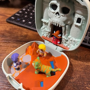 Op de afbeelding: Een miniatuur speelset met een schedelvormig kasteel. Binnenin bevinden zich verschillende kleine actiefiguren, waaronder een tijgerachtig wezen en andere personages. De speelset is voornamelijk oranje, grijs en groen.