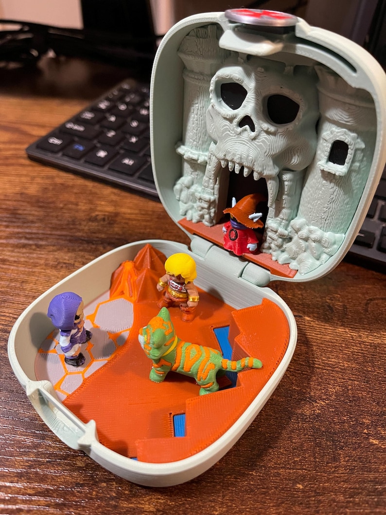 Op de afbeelding: Een miniatuur speelset met een schedelvormig kasteel. Binnenin bevinden zich verschillende kleine actiefiguren, waaronder een tijgerachtig wezen en andere personages. De speelset is voornamelijk oranje, grijs en groen.
