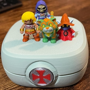 Op de afbeelding: Een lichtblauwe, afgeronde container met een rood en grijs embleem aan de voorkant, met vier miniatuur actiefiguren. De figuren zijn onder meer een blondharig personage, een groen-oranje tijger, een skeletfiguur en een rood gekleed personage.