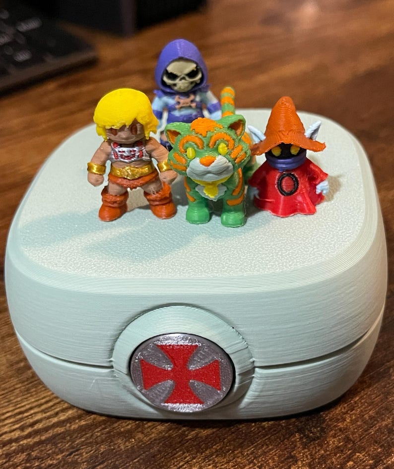 Op de afbeelding: Een lichtblauwe, afgeronde container met een rood en grijs embleem aan de voorkant, met vier miniatuur actiefiguren. De figuren zijn onder meer een blondharig personage, een groen-oranje tijger, een skeletfiguur en een rood gekleed personage.