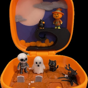 Op de afbeelding: Een oranje pompoenvormig speelgoed opent om een ​​Halloween-scène te onthullen. Binnenin zijn miniatuurfiguren van een skelet, een spook, een zwarte kat en een pompoenhoofdige vogelverschrikker. De achtergrond heeft een maan en pompoenen.