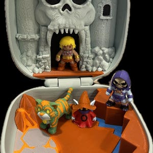 May include: A gray, hinged playset featuring a skull-shaped castle facade. Inside, miniature figures of a hero, a tiger, a wizard, and a skeletal villain are arranged on an orange and blue landscape. The playset is open, revealing the interior.