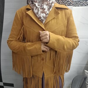 May include: A mustard yellow suede jacket featuring long fringe detailing along the chest, sleeves, and hemline. The jacket has a collared neckline, a button-front closure, and long sleeves. The material has a soft, matte texture throughout the garment.