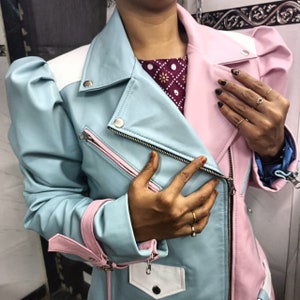 Peut inclure: Une veste en cuir bleu clair et rose avec une fermeture éclair et une poche blanche sur le devant. La veste a une bordure rose et un col blanc.