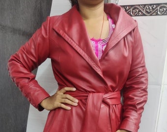 May include: A red leather trench coat with a wide collar, long sleeves, and a belted waist. The coat has a flared skirt and button details on the sleeves. The coat is knee-length and appears to be made of a smooth leather material.