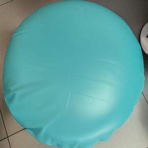 May include: A round, turquoise-colored seat cushion. The cushion has a smooth, slightly textured surface and appears to be made of a synthetic material. The cushion is set against a neutral-toned tiled floor.