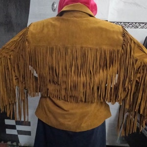 K&ouml;nnte beinhalten: Eine hellbraune Wildlederjacke mit langen, flie&szlig;enden Fransen am R&uuml;cken und an den &Auml;rmeln. Die Jacke hat einen klassischen Kragen und ein strukturiertes Passe-Design, wobei die Fransen von den Schultern bis zur R&uuml;ckenmitte reichen.