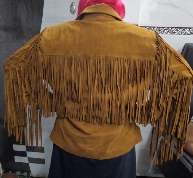 K&ouml;nnte beinhalten: Eine hellbraune Wildlederjacke mit langen, flie&szlig;enden Fransen am R&uuml;cken und an den &Auml;rmeln. Die Jacke hat einen klassischen Kragen und ein strukturiertes Passe-Design, wobei die Fransen von den Schultern bis zur R&uuml;ckenmitte reichen.