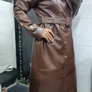May include: A long, dark brown leather trench coat featuring a double-breasted front with gold-toned buttons, a matching waist belt with a buckle, long sleeves with buttoned cuffs, and a vertical zippered pocket on the right side of the torso.