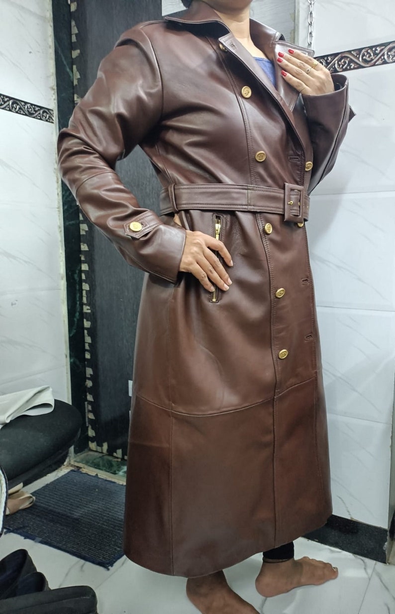 May include: A long, dark brown leather trench coat featuring a double-breasted front with gold-toned buttons, a matching waist belt with a buckle, long sleeves with buttoned cuffs, and a vertical zippered pocket on the right side of the torso.