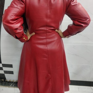 May include: A long-sleeved red coat featuring a structured collar, puffed shoulders, and a flared skirt. The coat has buttoned cuffs with gold-toned hardware and a smooth, solid finish, extending to a mid-calf length with a tailored back seam.