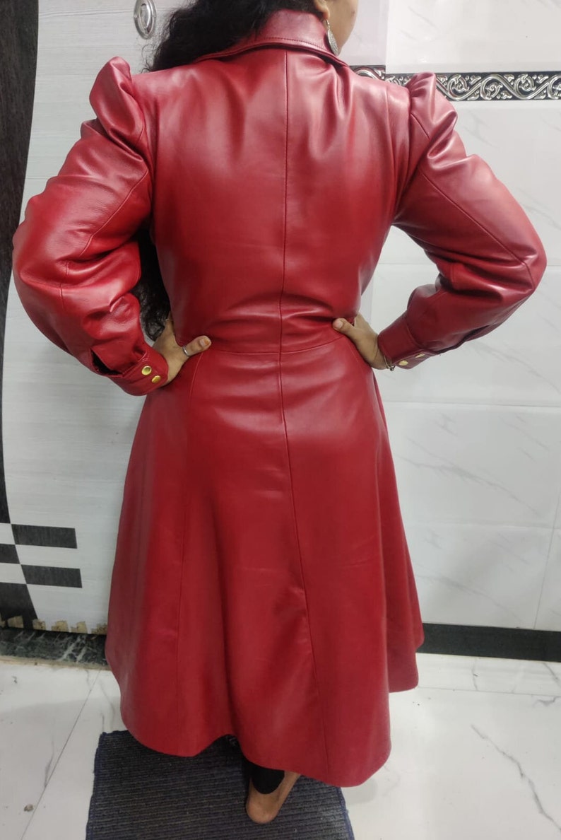 May include: A long-sleeved red coat featuring a structured collar, puffed shoulders, and a flared skirt. The coat has buttoned cuffs with gold-toned hardware and a smooth, solid finish, extending to a mid-calf length with a tailored back seam.