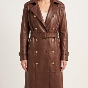 May include: A long, double-breasted brown leather trench coat featuring a wide notched collar, gold-toned buttons, and a matching belt with a rectangular buckle. The coat includes vertical zippered pockets on the chest and buckled sleeve straps.