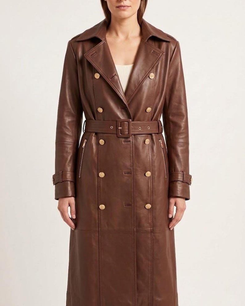 May include: A long, double-breasted brown leather trench coat featuring a wide notched collar, gold-toned buttons, and a matching belt with a rectangular buckle. The coat includes vertical zippered pockets on the chest and buckled sleeve straps.