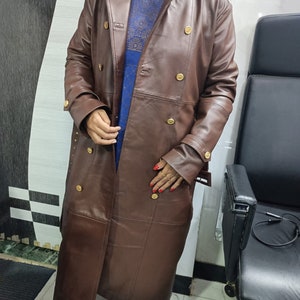 May include: A long, dark brown trench coat featuring a double-breasted front with gold-toned buttons, a wide collar, and long sleeves with buckled cuffs. The coat is worn over a blue patterned garment that is visible through the open front.