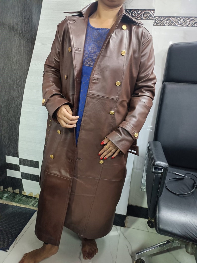 May include: A long, dark brown trench coat featuring a double-breasted front with gold-toned buttons, a wide collar, and long sleeves with buckled cuffs. The coat is worn over a blue patterned garment that is visible through the open front.