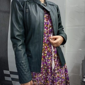 May include: A dark green jacket with a mandarin collar, a front zipper, and quilted detailing on the shoulders and cuffs. The jacket is worn over a purple, yellow, and white floral print dress featuring a white drawstring at the waist.