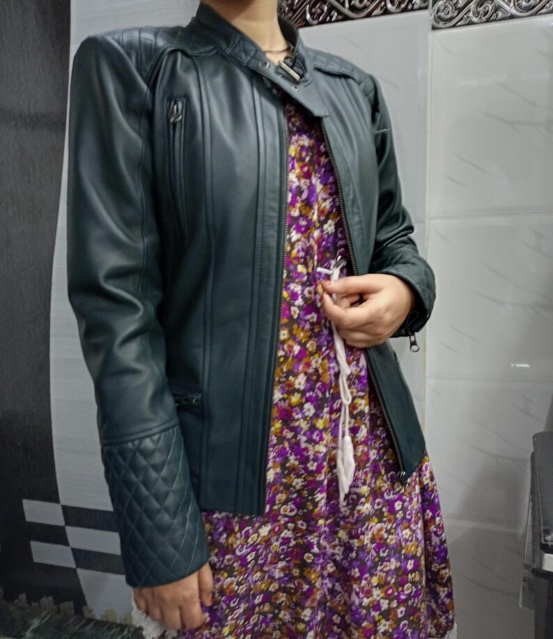 May include: A dark green jacket with a mandarin collar, a front zipper, and quilted detailing on the shoulders and cuffs. The jacket is worn over a purple, yellow, and white floral print dress featuring a white drawstring at the waist.