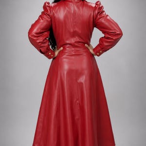 May include: A long-sleeved, floor-length red coat made of a smooth, leather-like material. The coat features a structured collar, puffed shoulders, and long sleeves with cuffs secured by gold-toned buttons. The garment has a fitted waist and a flared skirt.
