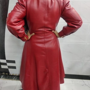 May include: A long-sleeved, knee-length red coat featuring a structured collar, puffed shoulders, and a fitted waistline. The coat is crafted from a smooth, solid red material and includes gold-toned buttons on the cuffs of the sleeves.