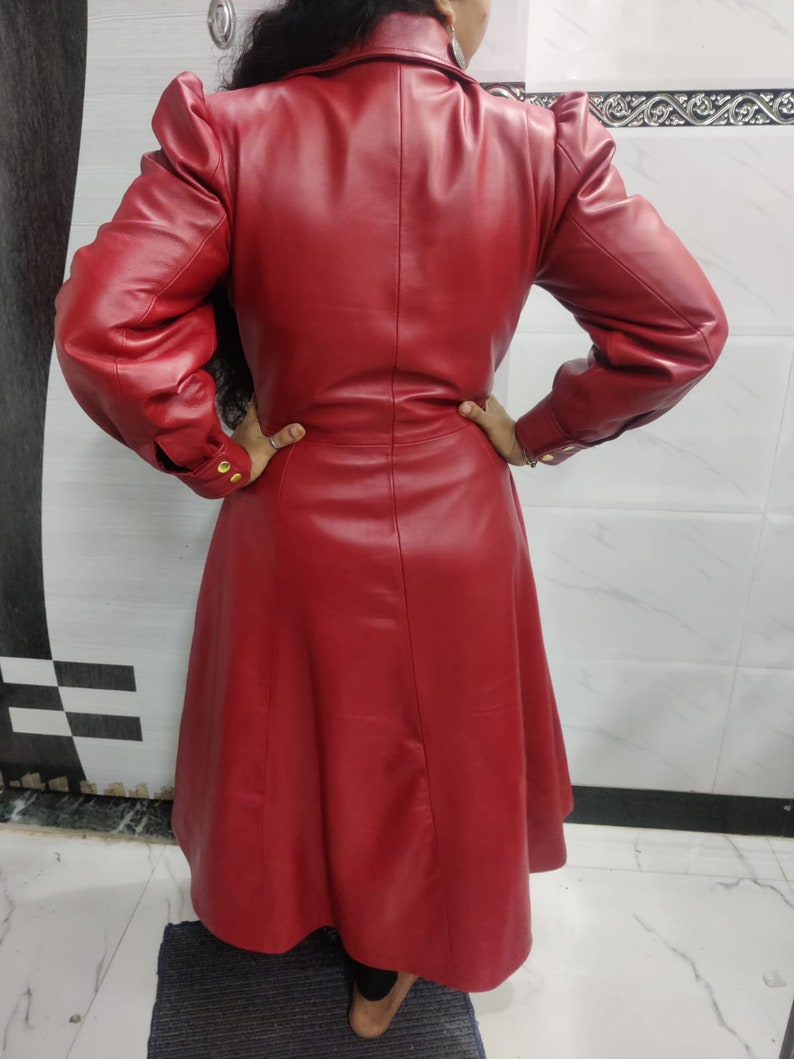 May include: A long-sleeved, knee-length red coat featuring a structured collar, puffed shoulders, and a fitted waistline. The coat is crafted from a smooth, solid red material and includes gold-toned buttons on the cuffs of the sleeves.