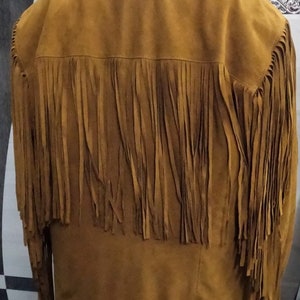 K&ouml;nnte beinhalten: Eine senffarbene Jacke mit klassischem Kragen und langen &Auml;rmeln. Das R&uuml;ckenteil und die &Auml;rmel sind mit langen, vertikalen Fransen verziert, die &uuml;ber die Schultern und die Arme verlaufen und f&uuml;r eine strukturierte Optik sorgen.