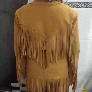 May include: A mustard yellow jacket featuring long fringe detailing across the back yoke and along the bottom hem. The jacket has a collared neckline and long sleeves, with the fringe extending from the shoulders down to the mid-back area.