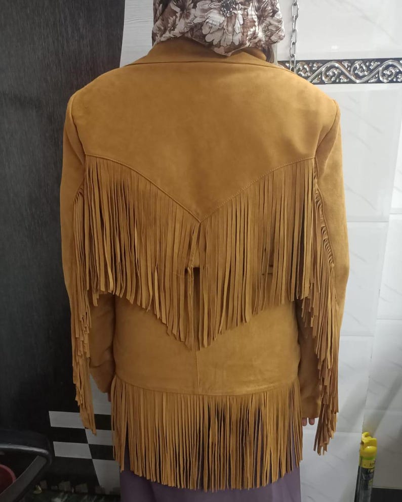 May include: A mustard yellow jacket featuring long fringe detailing across the back yoke and along the bottom hem. The jacket has a collared neckline and long sleeves, with the fringe extending from the shoulders down to the mid-back area.