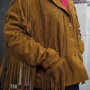 K&ouml;nnte beinhalten: Eine hellbraune Wildlederjacke mit langen Fransen an Brust, &Auml;rmeln und R&uuml;cken. Die Jacke verf&uuml;gt &uuml;ber einen klassischen Kragen, eine Knopfleiste mit drei sichtbaren braunen Kn&ouml;pfen und zwei Seitentaschen. Das Material hat eine matte Oberfl&auml;che.