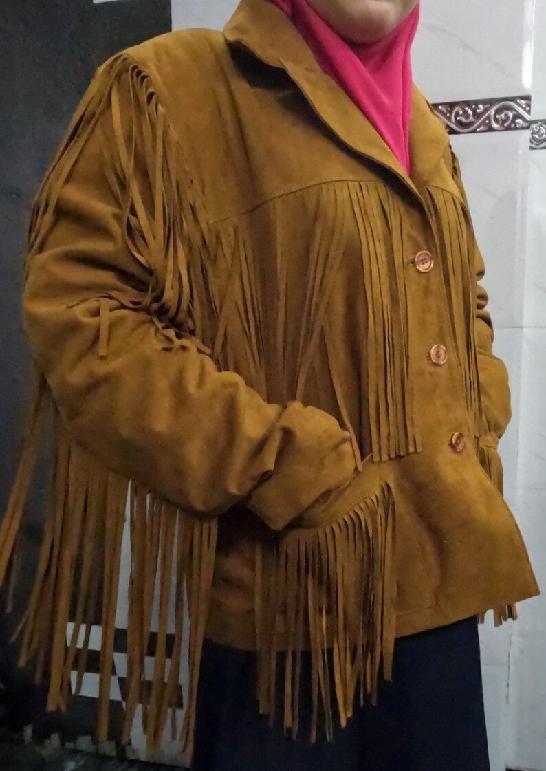 K&ouml;nnte beinhalten: Eine hellbraune Wildlederjacke mit langen Fransen an Brust, &Auml;rmeln und R&uuml;cken. Die Jacke verf&uuml;gt &uuml;ber einen klassischen Kragen, eine Knopfleiste mit drei sichtbaren braunen Kn&ouml;pfen und zwei Seitentaschen. Das Material hat eine matte Oberfl&auml;che.