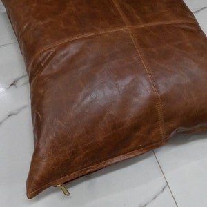 May include: A brown leather cushion with a zipper closure. The cushion is made of four panels of leather sewn together.