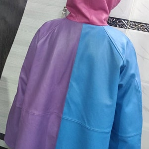 May include: A hooded jacket featuring a color-blocked design with a pink hood, a purple left panel, and a bright blue right panel. The jacket has long sleeves and a smooth, solid-colored finish, showcasing a distinct three-tone aesthetic.