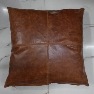 May include: A brown leather square cushion with a stitched design. The cushion is made of genuine leather and has a zipper closure.