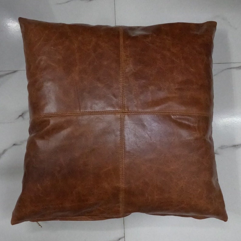 May include: A brown leather square cushion with a stitched design. The cushion is made of genuine leather and has a zipper closure.