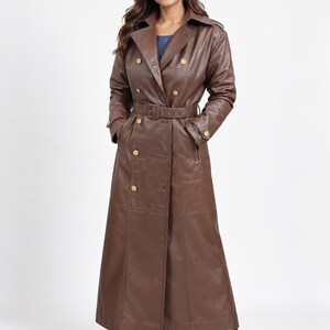 May include: A long, double-breasted brown leather trench coat featuring a wide collar, gold-toned buttons, and a matching belt with a buckle at the waist. The coat has long sleeves with buckled cuffs and reaches down to the ankles, paired with black boots.