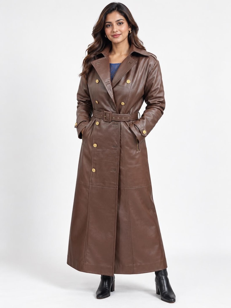 May include: A long, double-breasted brown leather trench coat featuring a wide collar, gold-toned buttons, and a matching belt with a buckle at the waist. The coat has long sleeves with buckled cuffs and reaches down to the ankles, paired with black boots.