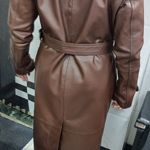 May include: A long, brown leather trench coat featuring a wide collar, long sleeves, and a matching belt cinched at the waist. The coat has a smooth finish, a central back vent, and structured shoulder detailing, extending down to the lower calf area.