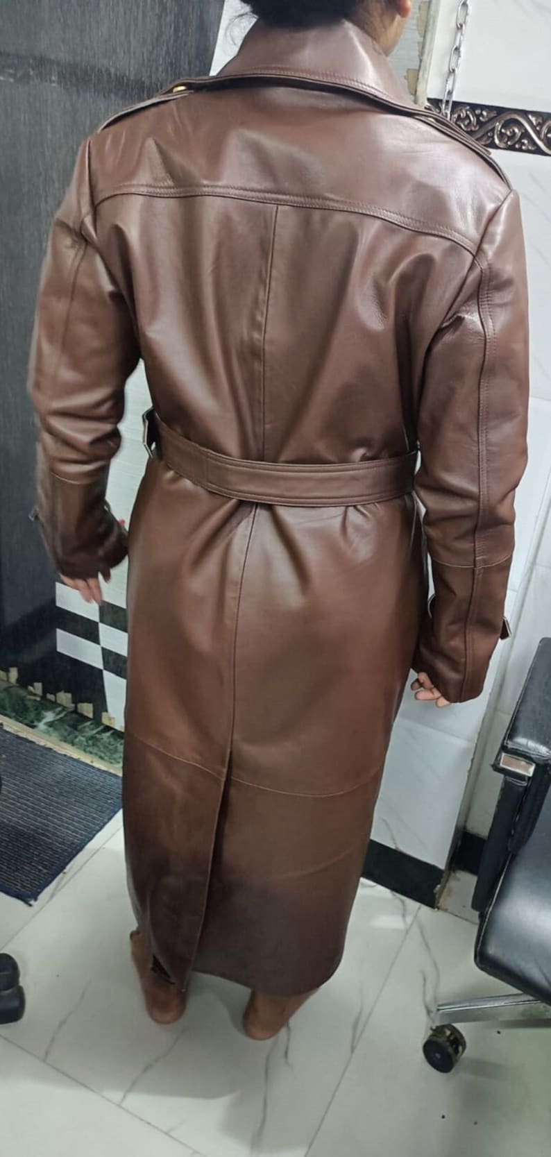 May include: A long, brown leather trench coat featuring a wide collar, long sleeves, and a matching belt cinched at the waist. The coat has a smooth finish, a central back vent, and structured shoulder detailing, extending down to the lower calf area.