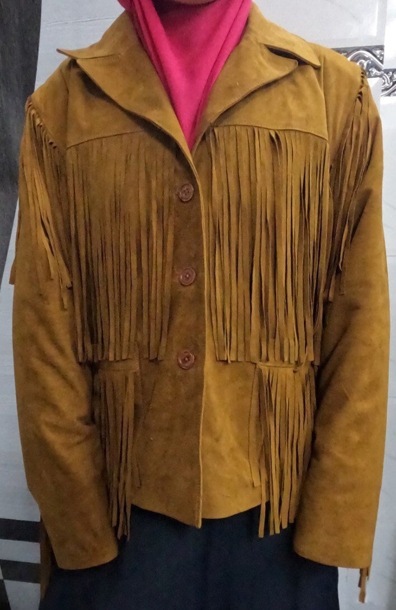 K&ouml;nnte beinhalten: Eine senfgelbe Jacke mit langen Fransen an der Brust und den unteren Vorderteilen. Die Jacke hat einen klassischen Kragen, lange &Auml;rmel und eine Knopfleiste mit drei passenden braunen Kn&ouml;pfen. Die Oberfl&auml;che wirkt weich und matt.