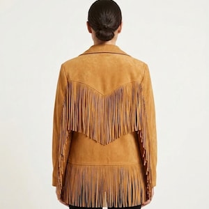 May include: A tan suede jacket featuring long fringe detailing across the back yoke and along the bottom hem. The jacket has a collared neckline and long sleeves, with the fringe creating a layered, textured appearance across the entire back.