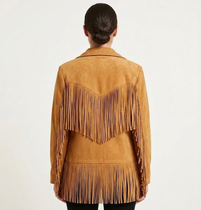 May include: A tan suede jacket featuring long fringe detailing across the back yoke and along the bottom hem. The jacket has a collared neckline and long sleeves, with the fringe creating a layered, textured appearance across the entire back.