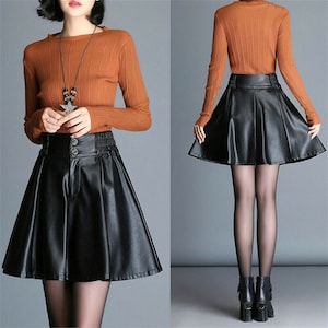 May include: A black faux leather skirt with a high waist and a flared silhouette. The skirt has a button closure and a pleated design. The model is wearing a brown sweater with a star-shaped pendant necklace.
