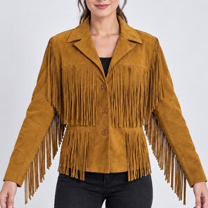 K&ouml;nnte beinhalten: Eine senfgelbe Wildlederjacke mit langen Fransen an Brust, &Auml;rmeln und unterem Saum. Die Jacke hat einen klassischen Kragen, eine Knopfleiste mit drei Kn&ouml;pfen und lange &Auml;rmel. Sie wird mit einer schwarzen Jeans kombiniert.