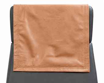 May include: A tan leather headrest cover is draped over the back of a black chair. The headrest cover is rectangular with stitched edges. The chair's back and seat are a dark gray color, creating a contrast with the tan cover.