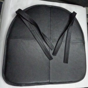 May include: A black, semi-circular seat cushion with four sections and attached straps. The cushion is made of a smooth, dark material, possibly leather or a leather-like fabric. The straps extend from the top of the cushion.