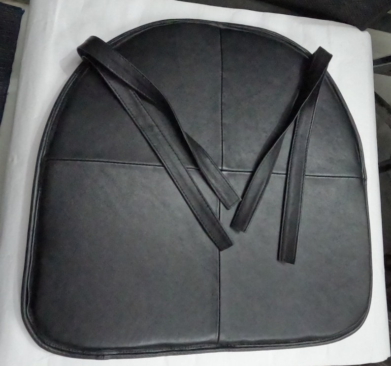 May include: A black, semi-circular seat cushion with four sections and attached straps. The cushion is made of a smooth, dark material, possibly leather or a leather-like fabric. The straps extend from the top of the cushion.