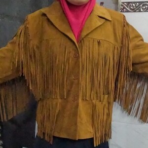 K&ouml;nnte beinhalten: Eine hellbraune Wildlederjacke mit langen Fransen an Brust, R&uuml;cken und &Auml;rmeln. Die Jacke verf&uuml;gt &uuml;ber einen klassischen Kragen, eine Knopfleiste auf der Vorderseite und lange &Auml;rmel. Das Material hat eine matte, strukturierte Wildlederoptik.