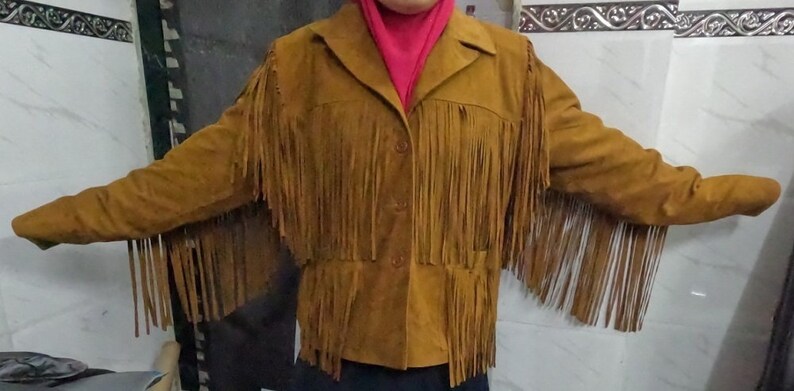 K&ouml;nnte beinhalten: Eine hellbraune Wildlederjacke mit langen Fransen an Brust, R&uuml;cken und &Auml;rmeln. Die Jacke verf&uuml;gt &uuml;ber einen klassischen Kragen, eine Knopfleiste auf der Vorderseite und lange &Auml;rmel. Das Material hat eine matte, strukturierte Wildlederoptik.