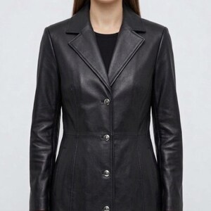 May include: A black leather coat featuring a notched lapel collar, long sleeves, and a front button closure with four silver-toned buttons. The coat has a tailored fit with visible vertical seam detailing along the front and a smooth finish.